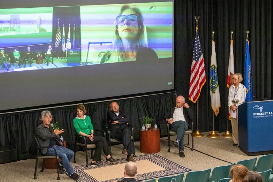 Natalie Roe (right, at the podium) moderates a discussion with panelists (seated from left to right) Hitoshi Murayama, Fabiolo Gianotti, Pier Oddone, and Michael Turner, with Maria Spiropulu engaging remotely via Zoom (onscreen, above right). (Credit: Robinson Kuntz, Berkeley Lab)