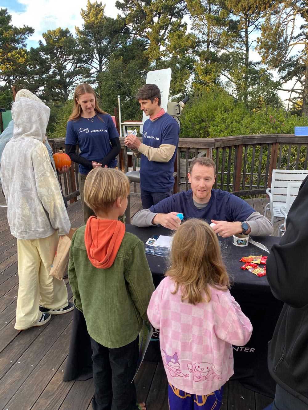 At left (background), Emily Perry (left) and Ibles Olcina talk with event participants while David Woodward (right, at the table in the foreground) sets up a detector demonstration with some of the younger participants. Credit: Daniel Kodroff, Berkeley Lab