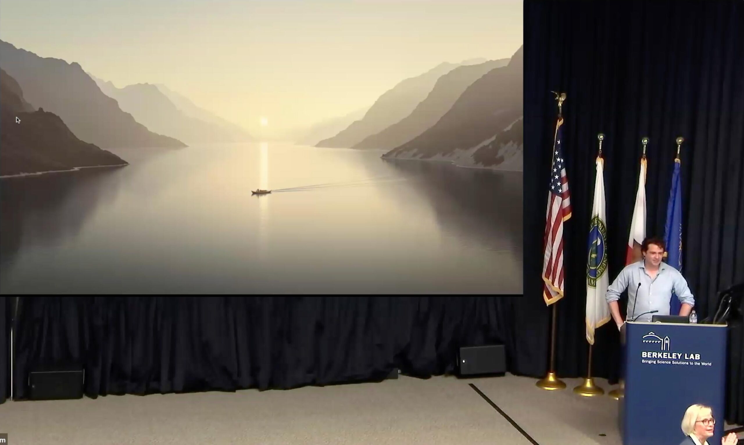 Ben Zevenbergen at the podium showing a slide with a landscape photo showing a large, flat waterway that reflects an overcast sky and the ridgelines of mountains on either side. In the midground is the silhouette of a person in a boat, sitting in the middle of the flat waterway that leads far away between fading mountains on either side and into a foggy sunrise/sunset.