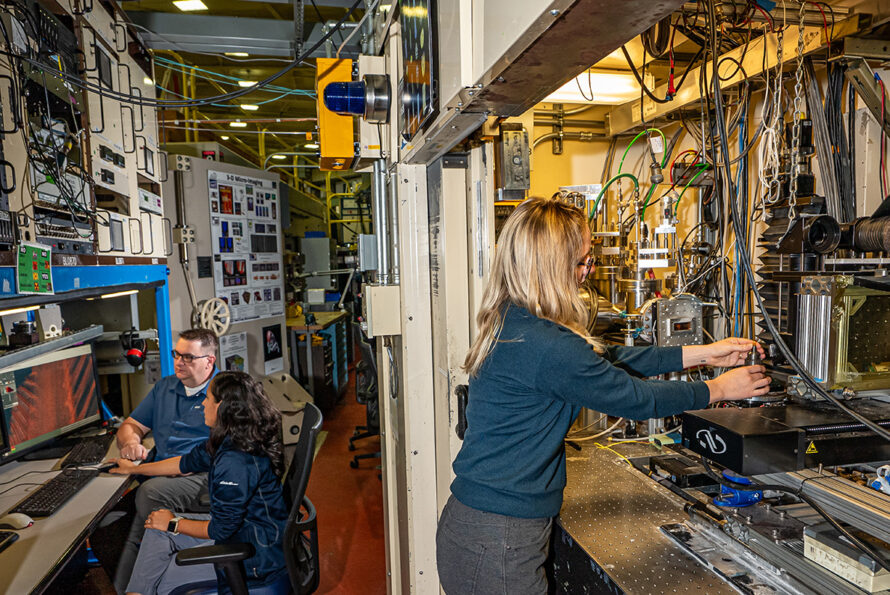 ALT: Two researchers in the background of a beamline laboratory look toward a computer with colorful microtomography imagery. A researcher is in the foreground making adjustments to an experimental setup.