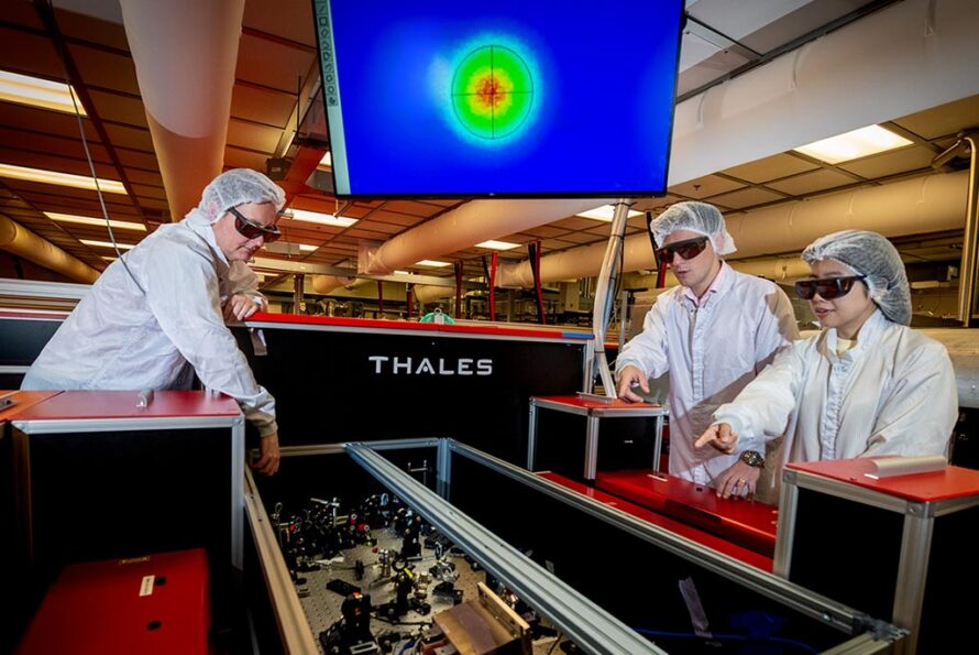 Three researchers in white coveralls and protective eyewear stand over equipment in a lab. A monitor with colorful figures is in the background.