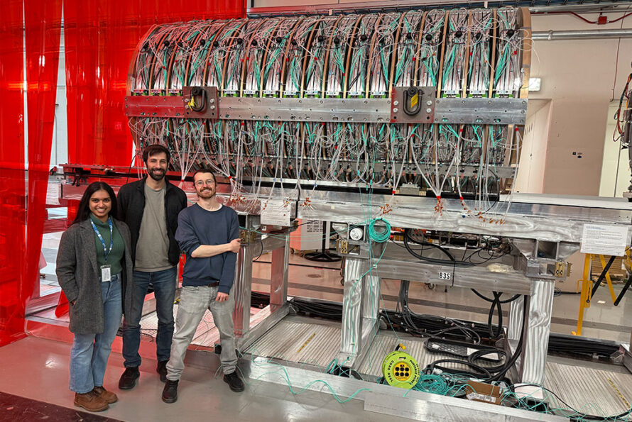 Namitha Chithirasreemadam, Richie Bonventre, and Ed Callaghan – members of Berkeley Lab’s Mu2e research team – in front of the Mu2e tracker, which is sitting on temporary support rails in the Mu2e experimental hall at Fermilab. (Photo credit: Vadim Rusu, Fermilab)