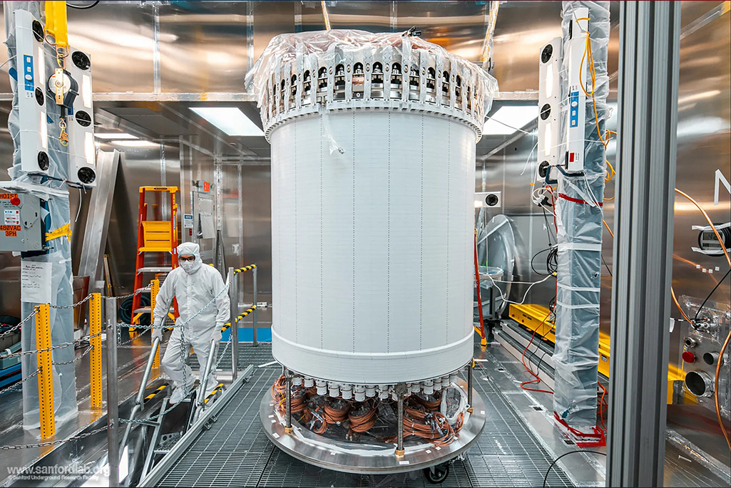 Caption: The LUX-ZEPLIN main detector in a surface lab before installation underground. Credit: Matthew Kapust/Sanford Underground Research Facility Alt text: A researcher in protective gear works near a tall, cylindrical scientific instrument with visible wiring. The walls surrounding it are reflective Teflon.