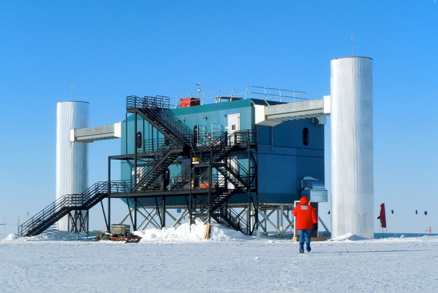 Alt Text: In the snow at the South Pole, a person in a bright red parka walks toward an enclosed, two-layered scientific facility with a system of criss-crossing black ladders on the side, and it's sitting on top of suspension girders and suspended between two (2) three-story-tall white towers.